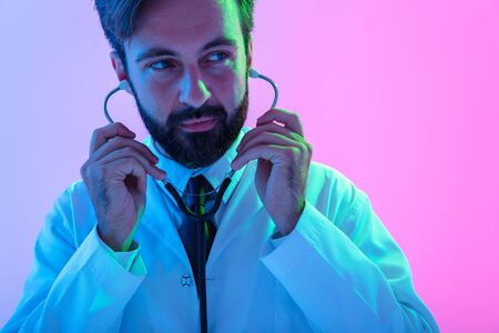 Portrait of a confident young man doctor wearing uniform standing isolated over pink-blue haze background, using stethoscopeの写真素材