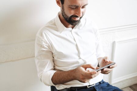 Image closeup of bearded pleased businessman wearing white shirt playing video game on cellphone...