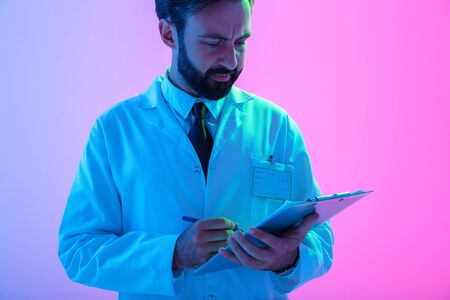 Cropped portrait of a confident young man doctor wearing uniform standing isolated over pink-blue haze background, taking notes in a notepadの写真素材