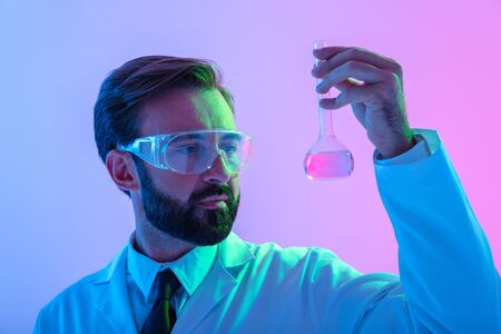 Portrait of a confident man scientist wearing unifrom and glasses standing isolated over blue-pink haze background? looking at a flask with liquidの写真素材
