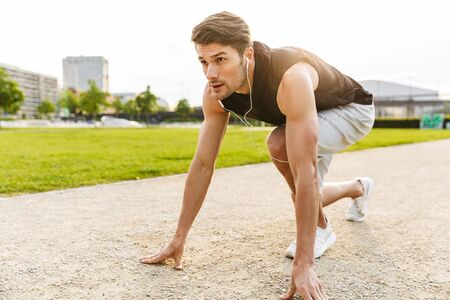 Image of athletic man in sportswear working out with earphones and running at green park outdoors in...