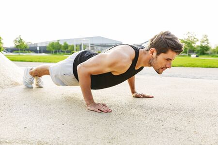 Image of handsome man in sportswear working out and doing push-ups at green park outdoors in morning