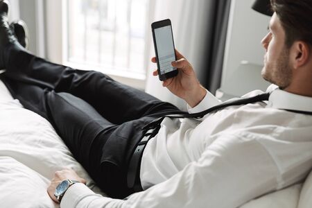 Image of unshaved young man wearing formal suit lying on bed with smartphone in hotel room during business tripの写真素材