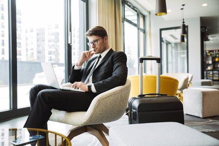 Portrait of beautiful young businessman wearing formal black suit sitting on armchair with laptop computer and suitcase in hotel hallの写真素材