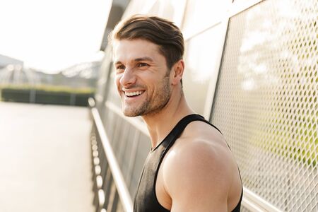 Photo of positive young man in sportswear smiling while standing on sports ground at green park in morningの写真素材