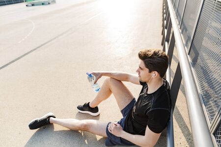 Portrait of handsome man in sportswear wearing earphones holding water bottle while sitting on...