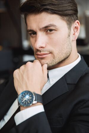 Portrait closeup of handsome young businessman wearing formal black suit posing with wristwatch indoorsの写真素材