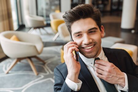 Photo of a young positive optimistic businessman indoors in business center office talking by mobile phone.の写真素材
