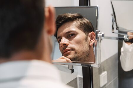 Image closeup of unshaved man wearing formal suit looking at mirror in bathroom at hotel room during business tripの写真素材