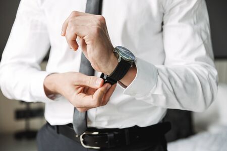 Cropped image of confident young man wearing formal suit taking off his wristwatch in hotel room...