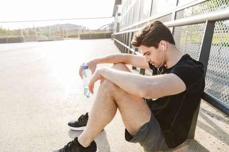 Portrait of brunette man in sportswear wearing earphones holding water bottle while sitting on...