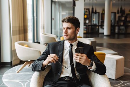 Photo of attractive young man wearing formal black suit sitting in hotel hall with smartphone and...