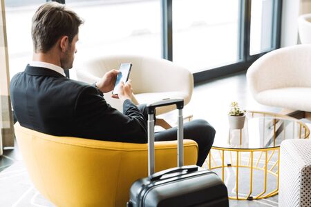 Photo of caucasian young man wearing formal black suit sitting in hotel hall with smartphone and suitcase during business tripの写真素材