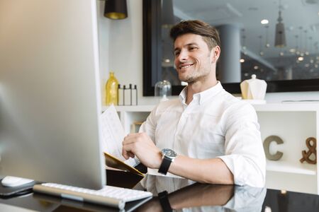 Image of smiling businesslike man wearing white shirt holding clipboard with papers while working on...