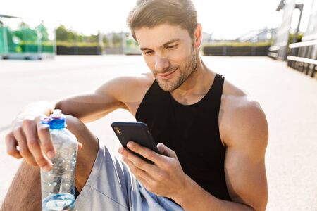 Portrait of young man in sportswear holding water bottle and cellphone while sitting on sports ground during morning workout outdoorsの写真素材