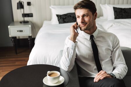 Image of handsome young man wearing formal suit talking on smartphone and drinking coffee in hotel apartment during business tripの写真素材