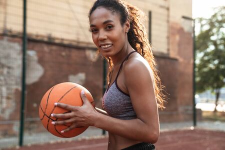 Image of joyful african american woman in sportswear playing basketball at playground outdoorsの写真素材