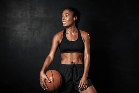 Image of smiling african american woman in sportswear holding basketball isolated over black...