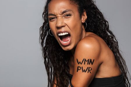 Portrait of a beautiful angry young african woman feminist standing isolated over gray background, screamingの写真素材