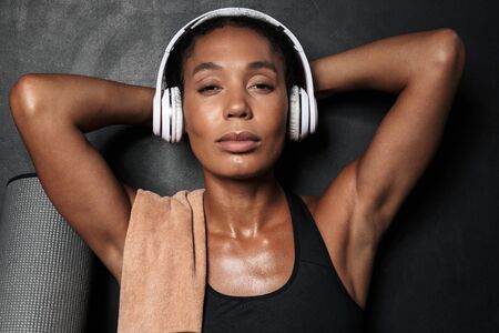 Attractive fit young athletic woman wearing sportswear sitting isolated over black background, listening to music with headphonesの写真素材