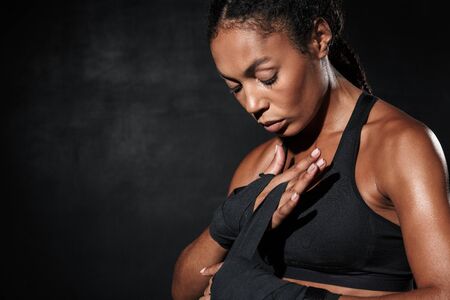 Image of athletic african american woman in sportswear wearing boxing hand wraps isolated over black backgroundの写真素材