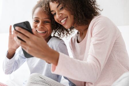 Image of pleased african american woman and her little daughter using cellphone together on sofa at homeの写真素材