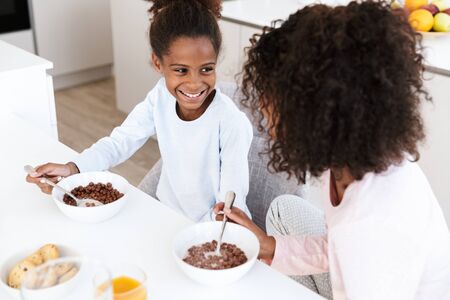 Image of african american woman and her little daughter having breakfast and eating corn flakes in kitchenの写真素材