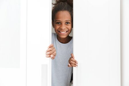 Image of cute african american little girl smiling and playing hide and seek in closet at homeの写真素材