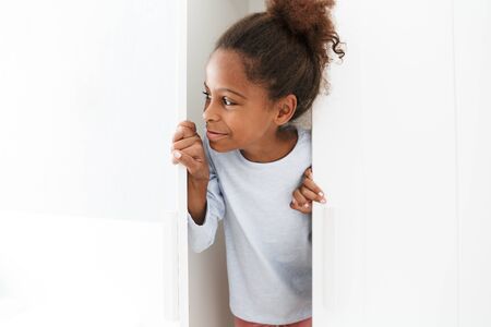 Image of attractive african american little girl smiling and playing hide and seek in closet at homeの写真素材