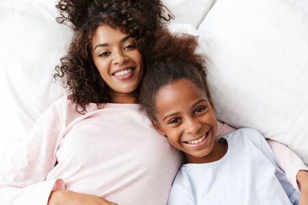 Image of cheerful african american woman and her daughter smiling and lying in bed at homeの写真素材