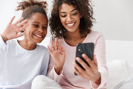 Image of cheerful african american woman and her little daughter using cellphone together on sofa at...