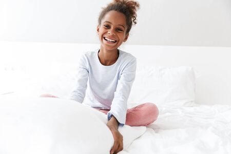 Image of satisfied african american little girl smiling and sitting in bed at homeの写真素材
