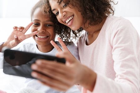 Image of joyful african american woman and her little daughter taking selfie photo on cellphone at homeの写真素材