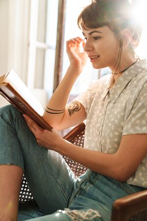 Photo of a young pretty woman indoors at home sit in chair reading book.