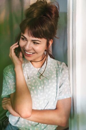 Image of a smiling pleased young beautiful woman talking by mobile phone indoors at home.の写真素材