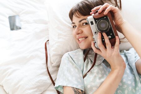Photo of a young pleased positive woman indoors at home lies on bed in the morning holding camera photographing.の写真素材