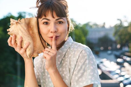 Photo of a positive happy young beautiful woman on a balcony holding shell near ear showing silence gesture.の写真素材