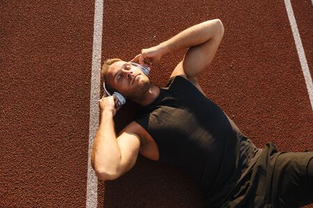 Image of athletic sportsman listening to music with headphones while lying at running track outdoorsの写真素材