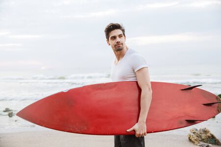 Image of a serious handsome young man surfer with surfing on a beach outside.