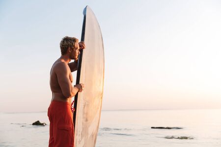 Image of blonde shirtless athletic surfer man holding his surfboard by ocean at sunriseの写真素材