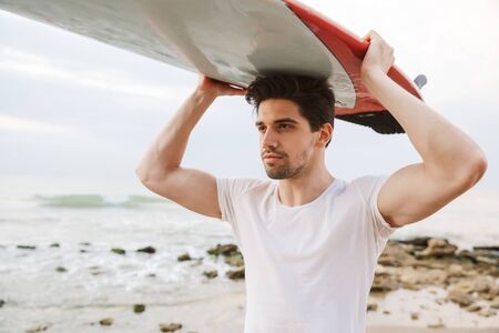 Photo of a serious concentrated handsome man surfer with surfing on a beach outside.
