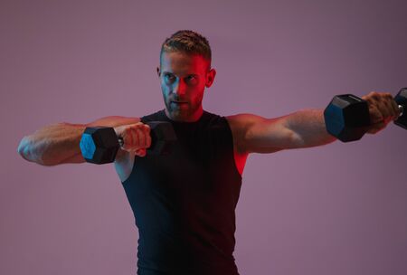Image of a concentrated young sports man posing isolated over neon lights make exercises with...
