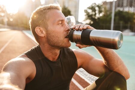 Image of muscular sportsman drinking water while doing workout at stadium outdoorsの写真素材