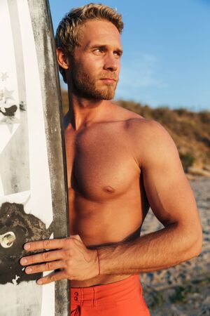 Image of muscular shirtless blonde surfer man holding his surfboard by ocean at sunriseの写真素材