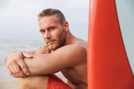 Image of a cheery positive strong handsome man surfer with surfing on a beach outside.の写真素材