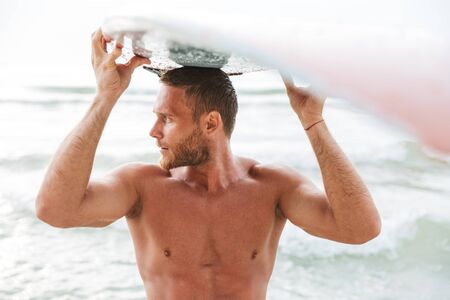 Image of a serious strong handsome man surfer with surfing on a beach outside.の写真素材