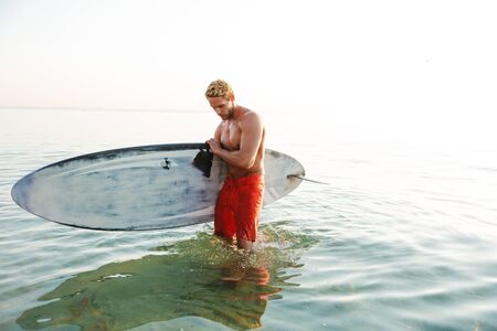 Picture of attractive concentrated young man surfer with surfing desk at the beach sea.