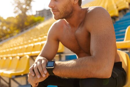 Image of masculine shirtless sportsman sitting on stadium seat during workout outdoors on sunny dayの写真素材