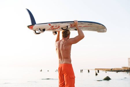 Image of strong young shirtless surfer man carrying his surfboard by ocean at sunrise