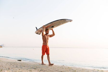 Image of muscular young shirtless surfer man carrying his surfboard by ocean at sunriseの写真素材